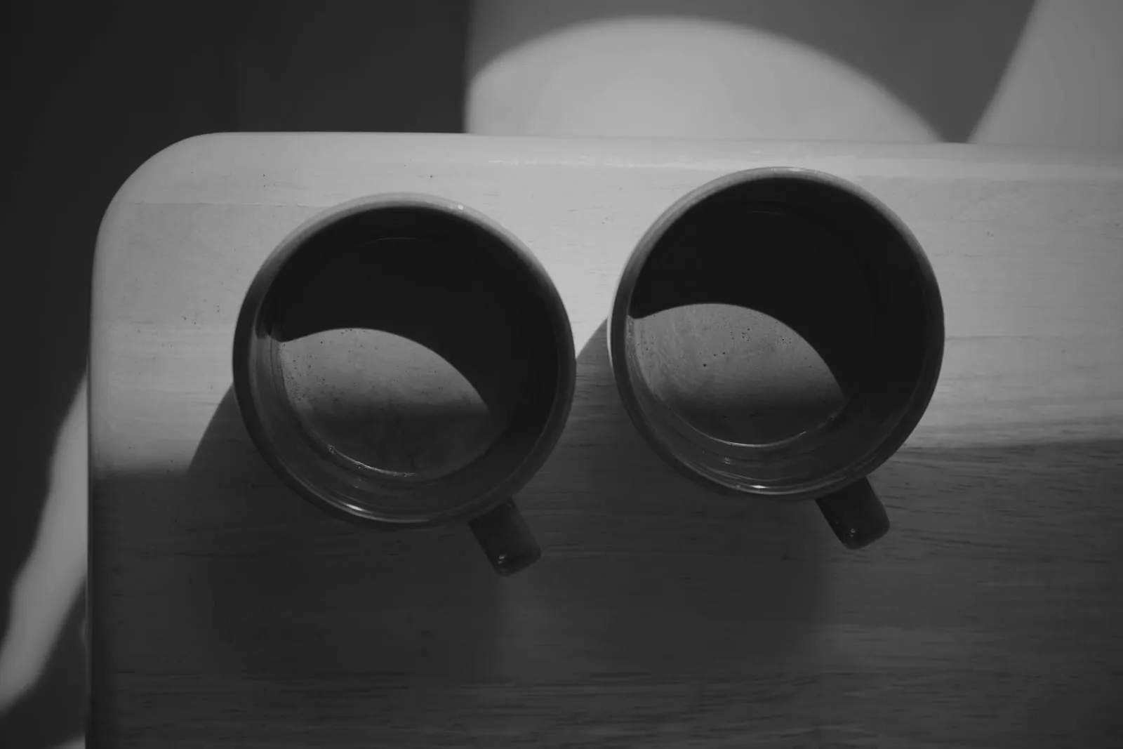 Zwei Kaffeebecher nebeneinander auf einem Holztisch, von oben, im Streiflicht der Morgensonne.