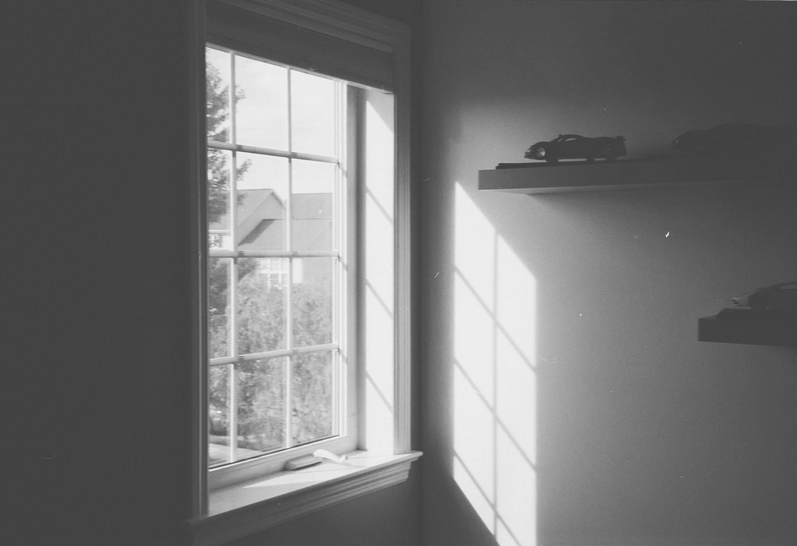 Ein Fenster mit Sprossen, das Morgenlicht wirft ein Fensterkreuz an die Wand, darüber ein Regal mit einem Spielzeugauto.